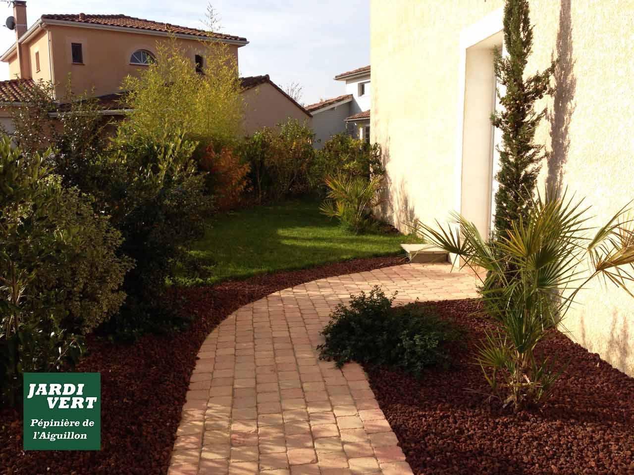 Jardin paysager avec allée pavée incurvée, paillis rouge. Plantes méditerranéennes, cyprès et maison. Jardi Vert Pépinière de l'Aiguillon.