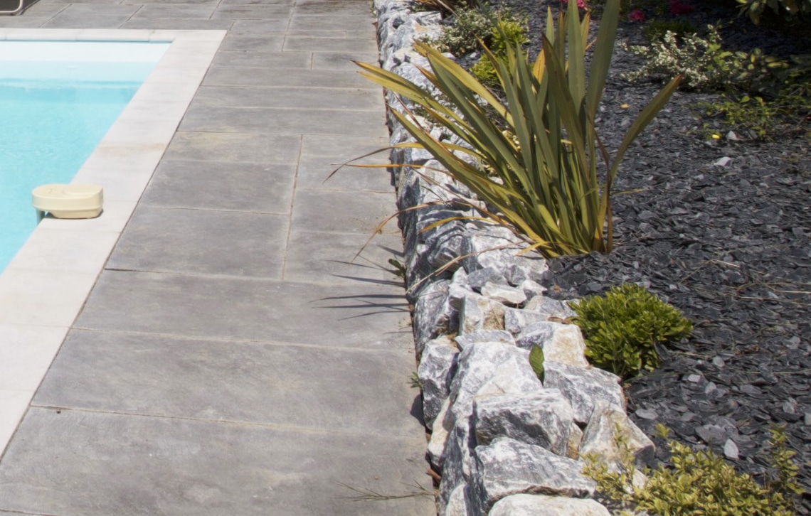 Terrasse en dalles grises longeant une piscine avec flotteur jaune. Muret de pierres et jardin paysager avec paillis d'ardoise et plantes.