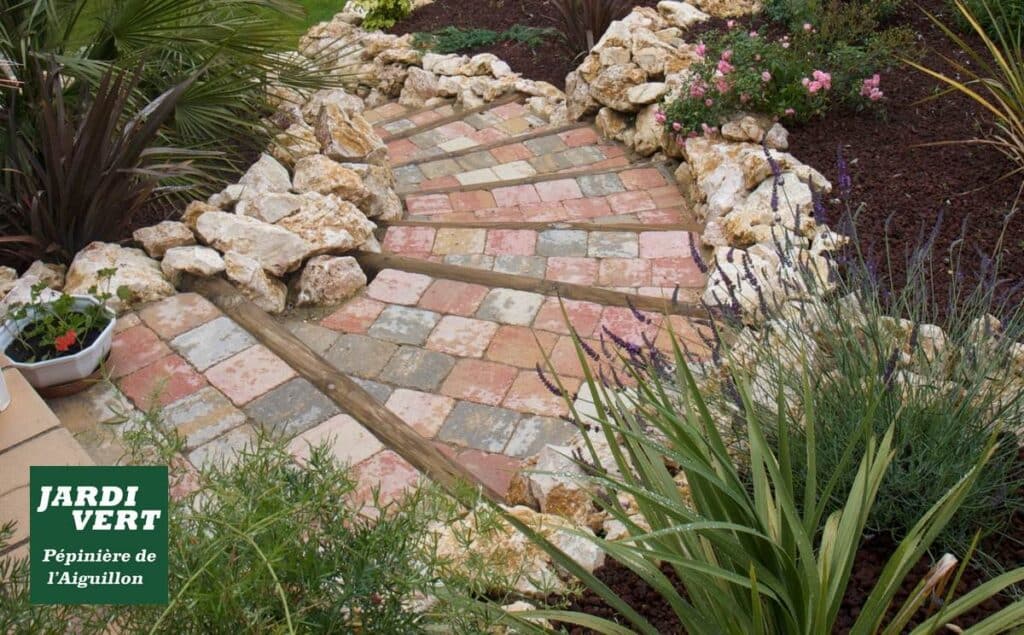 Allée de jardin en pavés multicolores avec marches incurvées, bordée de rochers et de plantes (lavande, roses). Logo : JARDI VERT Pépinière de l'Aiguillon.