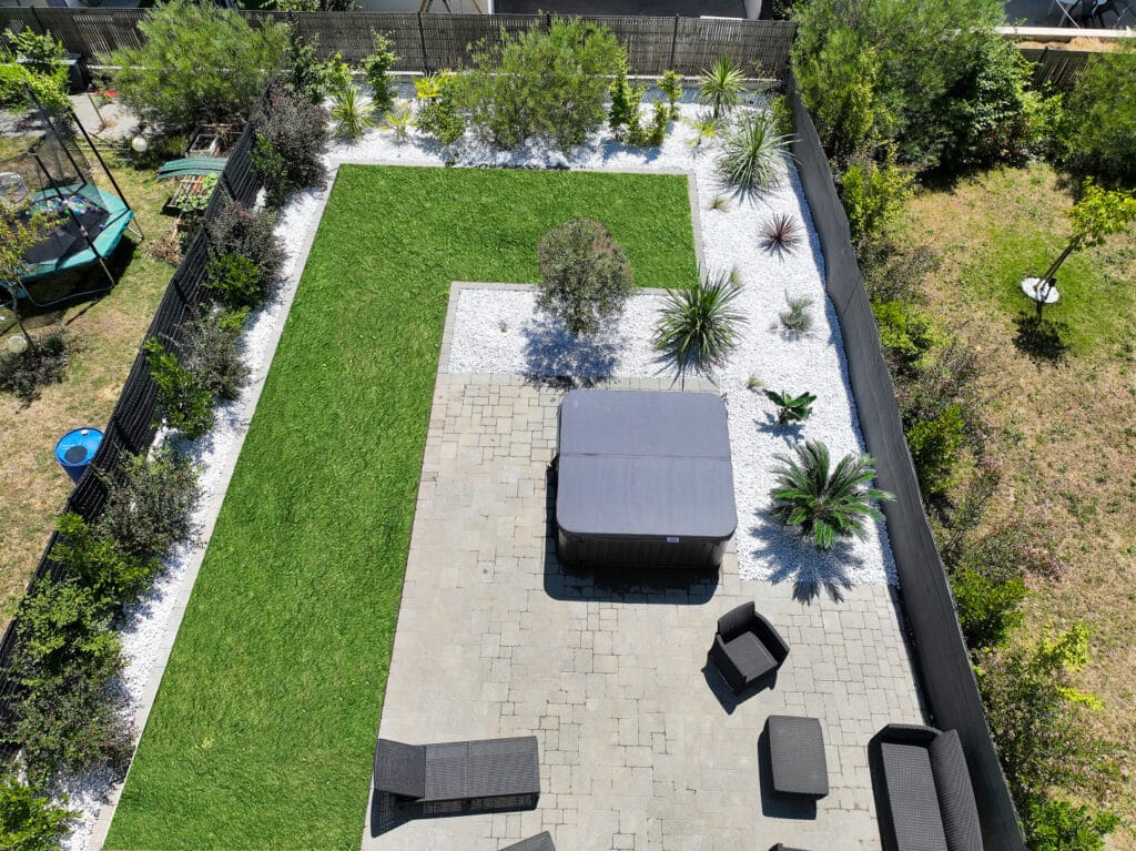 Jardin contemporain zen avec pelouse verte, galets blancs, plantes méditerranéennes, jacuzzi et mobilier sur terrasse pavée.