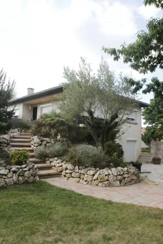 Maison moderne claire avec toit gris, volets blancs. Jardin méditerranéen en terrasses, olivier, murs de pierre, escalier en bois, pelouse.