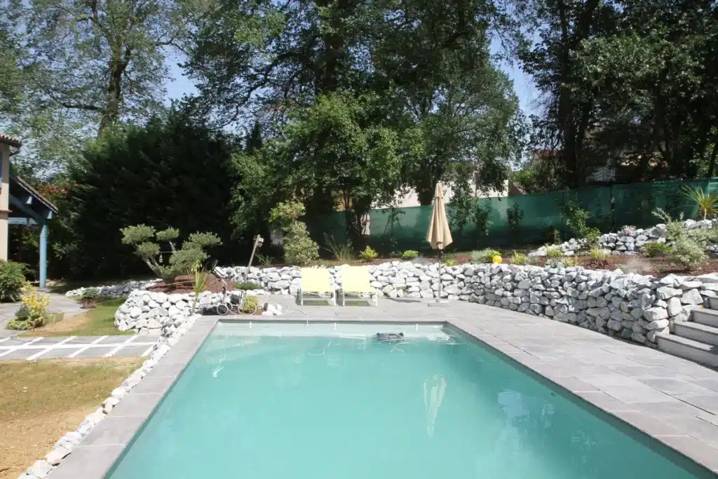 Piscine moderne avec terrasse pavée, mur de pierres sèches et jardin paysagé. Deux transats jaunes sous un parasol, arbres luxuriants.