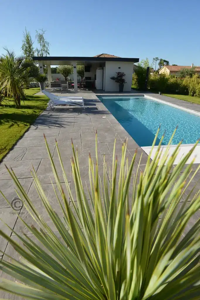 Piscine moderne rectangulaire et terrasse grise avec transat et pool house équipé, entourées de pelouse verdoyante sous un ciel bleu dégagé.