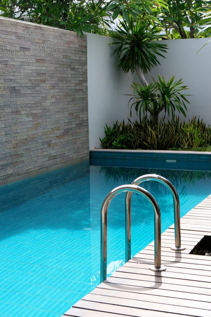 Piscine moderne turquoise avec échelle chromée et terrasse en bois. Un mur en pierre texturé et une végétation luxuriante bordent le bassin.