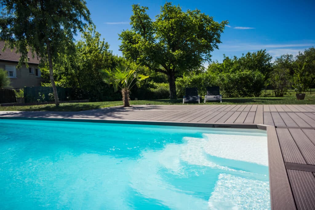 Piscine moderne avec eau turquoise, terrasse en bois, jardin verdoyant et chaises longues sous un ciel bleu ensoleillé.