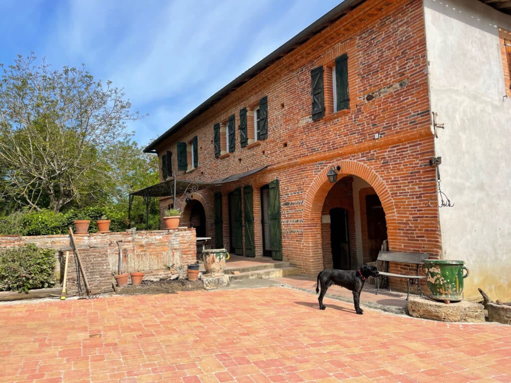 Maison ancienne en briques rouges typique du sud de la France avec volets verts, chiot noir sur terrasse dallée de terre cuite, outils de jardinage à côté d'un muret.