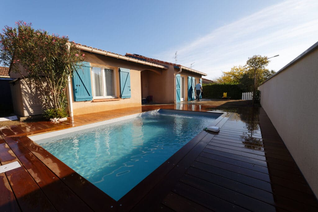 Maison provençale avec volets bleus et piscine rectangulaire neuve entourée d'une terrasse en bois foncé mouillée par temps clair.