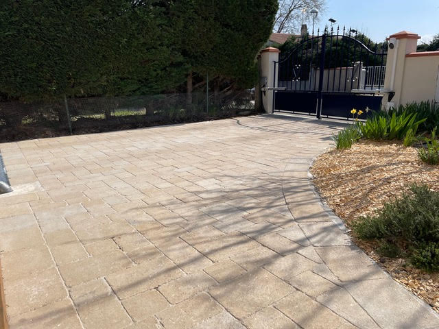 Allée pavée en pierre naturelle menant à un portail noir en fer forgé avec haie de troènes et jardin paysager.
