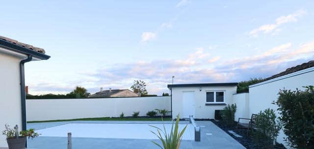 Piscine moderne avec volet roulant blanc, terrasse en dalles grises et pool house blanc sous un ciel bleu dégagé.